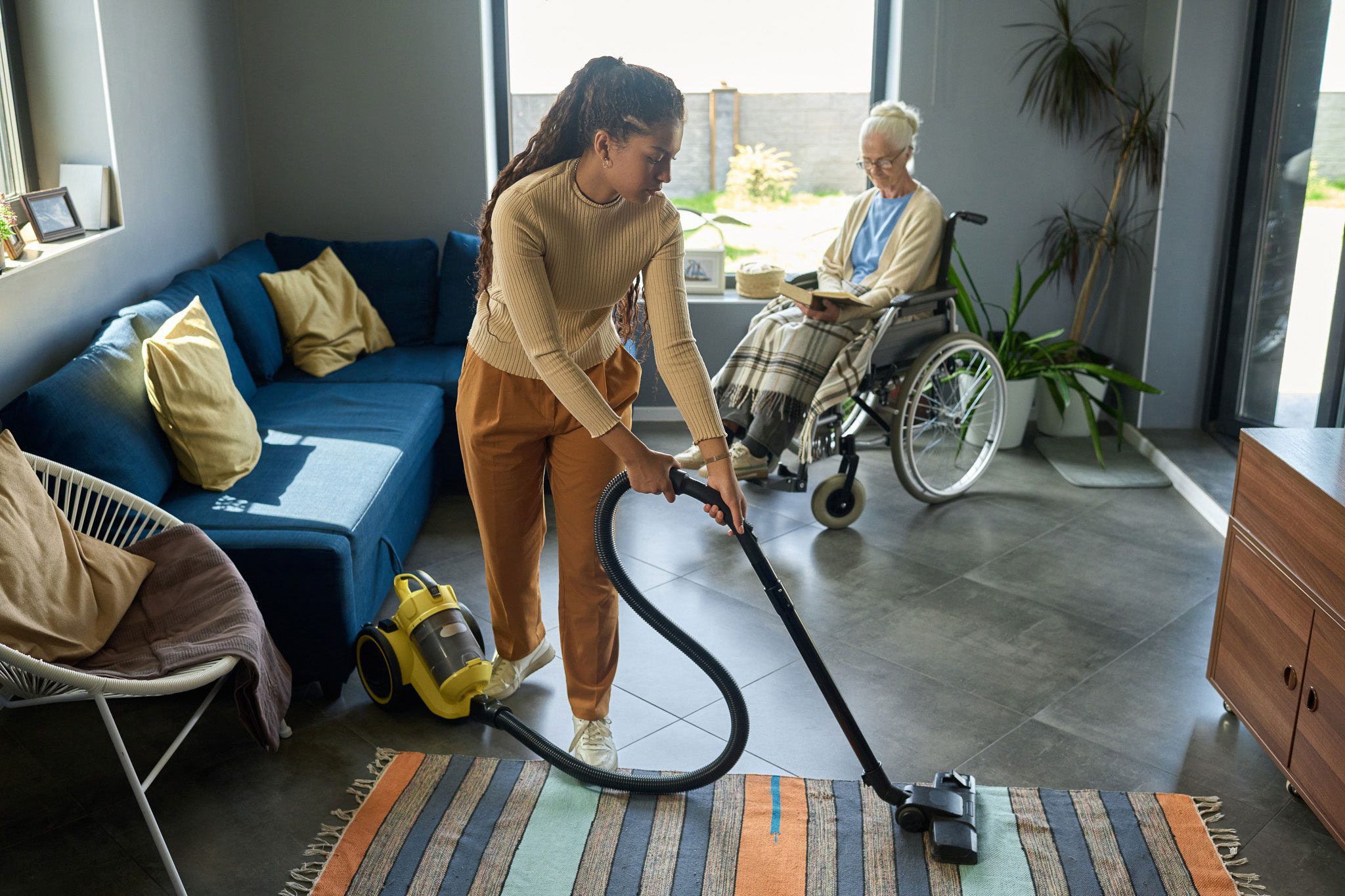A woman vacuums a rug in a living room while an older person in a wheelchair sits nearby by the window.