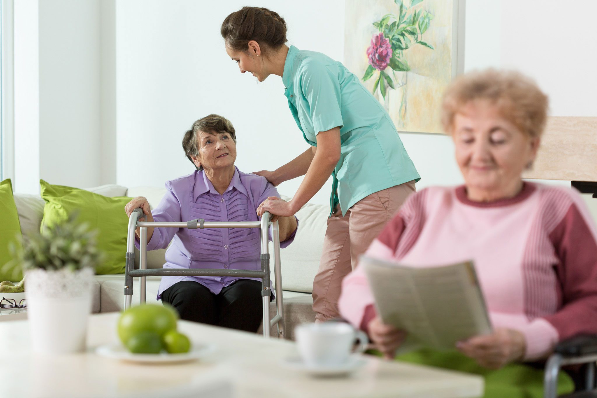 A caregiver assists an elderly woman using a walker while another elderly woman sits in the foreground reading a newspaper in a bright living room.